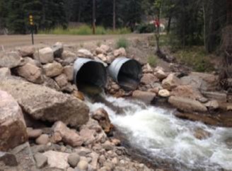 A photo showing two culverts with flowing water, surrounded by a rock embankment. The water is flowing through the culverts, and the rocks along the sides help support the structure. The scene is natural, with greenery visible in the background.