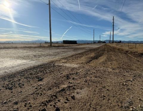 A rural scene showing a dirt shoulder along County Road 50, with a clear blue sky and light clouds overhead.