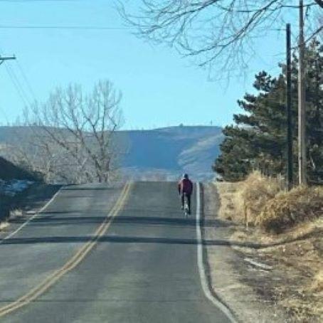 A cyclist riding up a hilly road with a clear blue sky and mountains in the background.